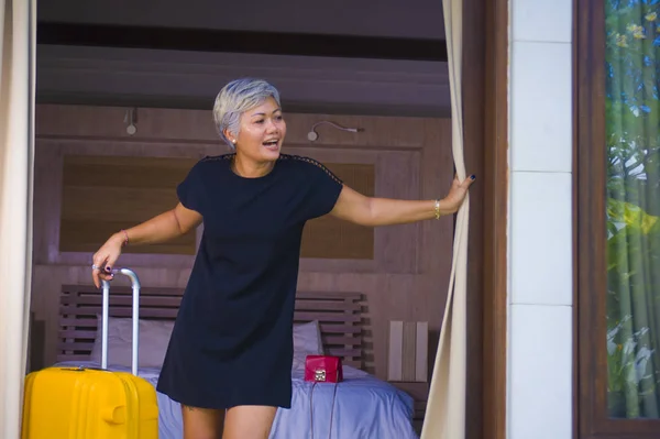 Привлекательная и счастливая азиатка средних лет в стильном летнем платье, прибывающая в гостиницу, открывающая дверь балкона, выглядящая радостно, наслаждаясь отпуском в отпуске — стоковое фото