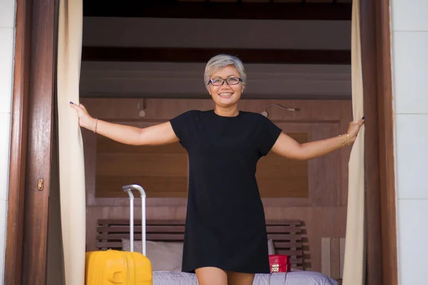 Привлекательная и счастливая азиатка средних лет в стильном летнем платье, прибывающая в гостиницу, открывающая дверь балкона, выглядящая радостно, наслаждаясь отпуском в отпуске — стоковое фото