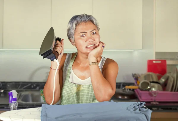 Attractive and stressed Asian middle aged lady ironing at home kitchen desperate and overwhelmed feeling unhappy in helpless face expression