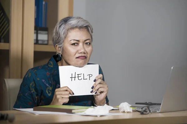Verzweifelte und gestresste attraktive asiatische Frau mittleren Alters, die einen Notizblock in der Hand hält und um Hilfe bittet, fühlt sich überarbeitet und ausgebeutet, wenn sie am Schreibtisch am Computer arbeitet und unter Stress leidet — Stockfoto