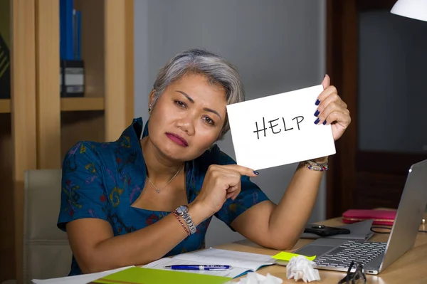 絶望的でストレスの多い魅力的な中年アジアの女性が、過労とストレスに苦しむコンピュータオフィスデスクで働くことを助けを求めるメモ帳を持っている — ストック写真