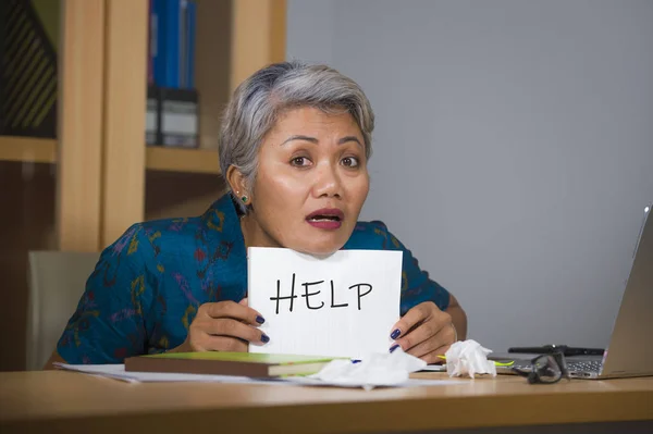 Verzweifelte und gestresste attraktive asiatische Frau mittleren Alters, die einen Notizblock in der Hand hält und um Hilfe bittet, fühlt sich überarbeitet und ausgebeutet, wenn sie am Schreibtisch am Computer arbeitet und unter Stress leidet — Stockfoto