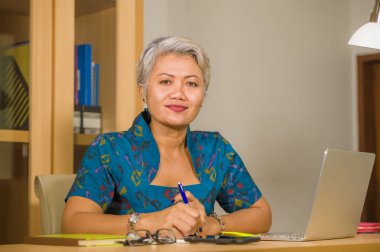 finansal iş başarı zarif elbise giyen emin gülümseyerek dizüstü bilgisayar masasında çalışan çekici ve mutlu başarılı orta yaşlı Asyalı kadın