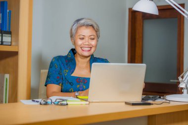 finansal iş başarı zarif elbise giyen emin gülümseyerek dizüstü bilgisayar masasında çalışan çekici ve mutlu başarılı orta yaşlı Asyalı kadın