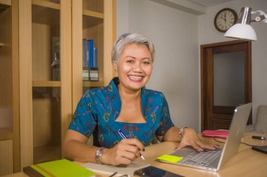 finansal iş başarı zarif elbise giyen emin gülümseyerek dizüstü bilgisayar masasında çalışan çekici ve mutlu başarılı orta yaşlı Asyalı kadın