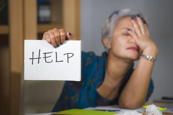 Desesperada y estresada atractiva mujer asiática de mediana edad sosteniendo bloc de notas pidiendo ayuda sintiéndose sobrecargada de trabajo y explotada trabajando en la oficina de informática sufriendo estrés Imagen De Stock