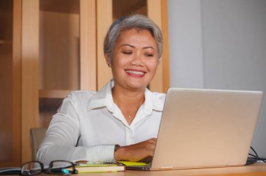 ofis dizüstü bilgisayar masasında çalışan mutlu ve başarılı çekici orta yaşlı Asyalı kadının kurumsal iş yaşam tarzı portre memnun ve iş başarısı verimli