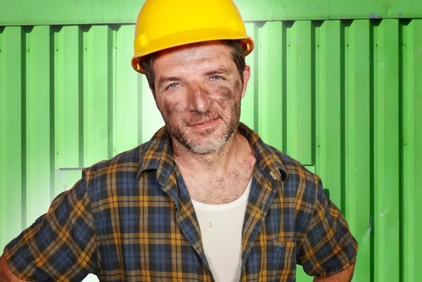 Junger attraktiver und selbstbewusster Bauunternehmer oder Bauarbeiter mit Bauarbeiterhelm posiert fröhlich lächelnd mit schmutzbeflecktem Gesicht auf der Baustelle — Stockfoto