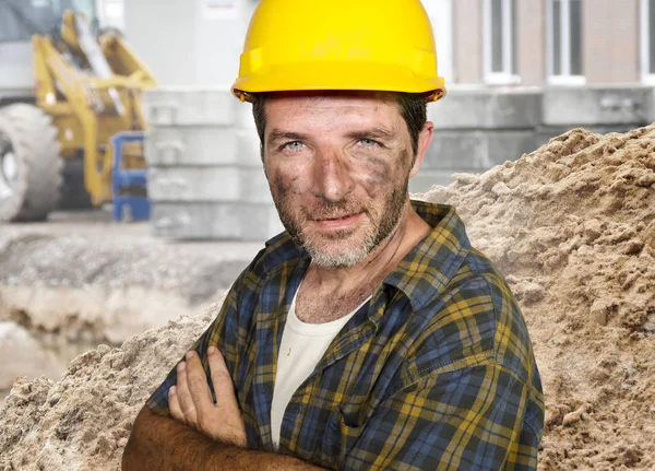 young attractive and confident contractor or construction worker man with builder safety helmet posing corporate smiling cheerful with dirt stained face at building site