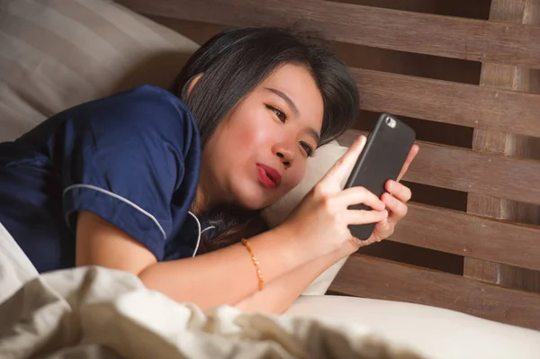 Joven hermosa y feliz mujer coreana asiática en pijama utilizando el teléfono móvil mensajes de texto en las redes sociales con su novio o disfrutar en línea citas aplicación sonriente alegre — Foto de Stock
