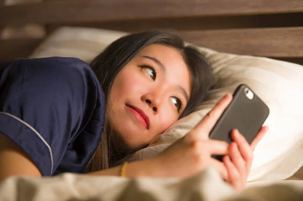 Joven hermosa y feliz mujer coreana asiática en pijama utilizando el teléfono móvil mensajes de texto en las redes sociales con su novio o disfrutar en línea citas aplicación sonriente alegre — Foto de Stock