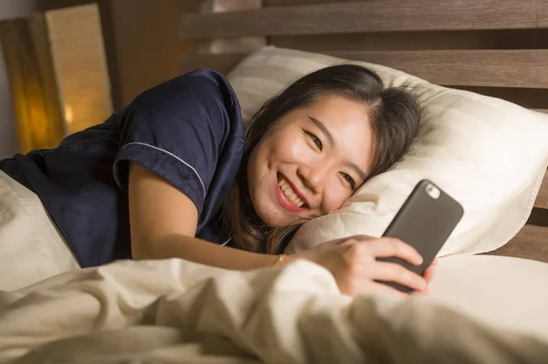 Молодая красивая и счастливая азиатская японка в пижаме с помощью мобильного телефона, смс-ки со своим парнем или приложение для онлайн знакомств, весело улыбающееся — стоковое фото