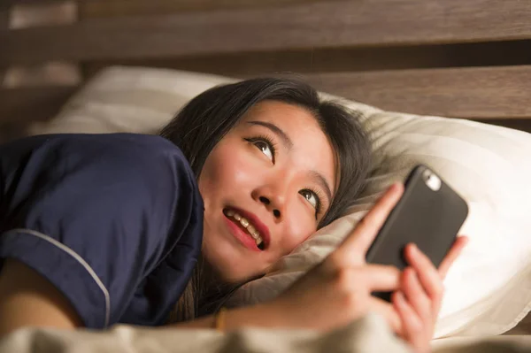 Jonge mooie en gelukkige Aziatische Japanse vrouw in pyjama met behulp van mobiele telefoon sociale media sms'en met haar vriendje of genieten van online dating app glimlachend vrolijk — Stockfoto