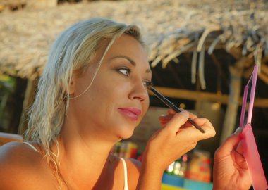 genç güzel ve rahat sarışın kadın moda ve güzellik konsepti tatil plaj beldesinde oturan fırça göz farı uygulayarak makyaj rötuş ayna tutan