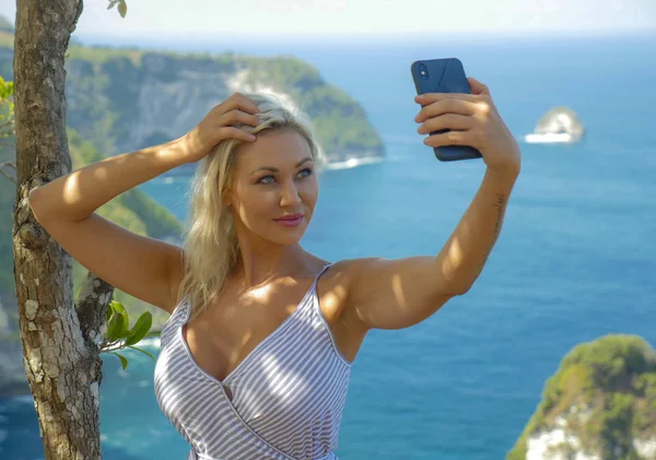 Junge glückliche und attraktive blonde Frau macht Selfie-Porträt mit Handy am schönen tropischen Paradies Blick auf Ocean Rock Cliff genießen Sommerferien — Stockfoto