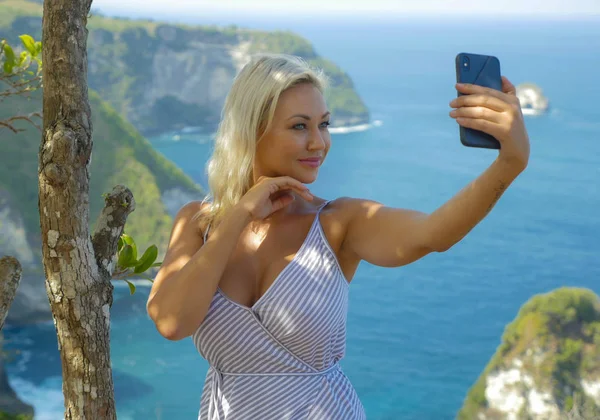 Молодая счастливая и привлекательная блондинка делает селфи-портрет с мобильным телефоном в красивом тропическом раю вид на скалу океана наслаждаясь летними каникулами — стоковое фото