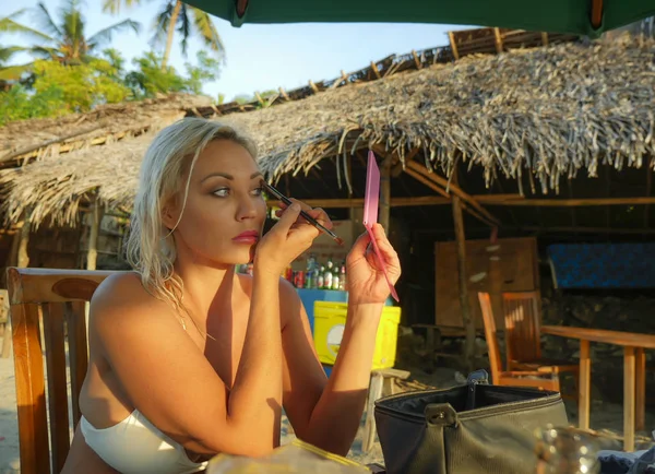Junge schöne und entspannte blonde Frau mit Spiegel retuschieren Make-up Anwendung Pinsel Lidschatten sitzen im Urlaub Strand Resort in Mode und Beauty-Konzept — Stockfoto