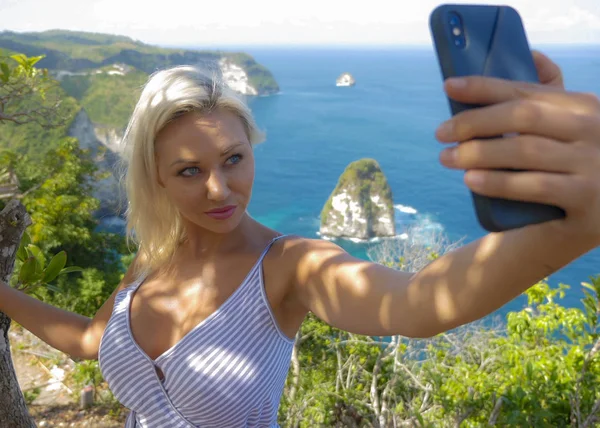 Junge glückliche und attraktive blonde Frau macht Selfie-Porträt mit Handy am schönen tropischen Paradies Blick auf Ocean Rock Cliff genießen Sommerferien — Stockfoto