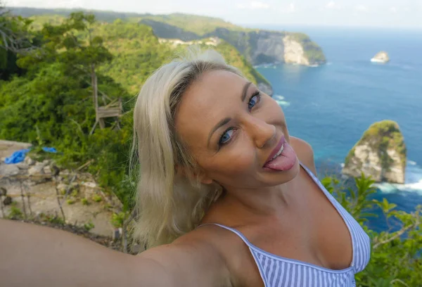 Молодая счастливая и привлекательная блондинка делает селфи-портрет с мобильным телефоном в красивом тропическом раю вид на скалу океана наслаждаясь летними каникулами — стоковое фото