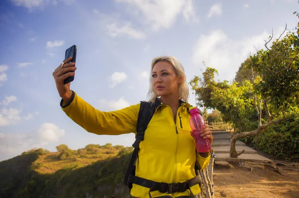 young beautiful and happy hiker woman with backpack hiking on sea mountain cliff taking selfie portrait with mobile phone enjoying solo travel trekking adventure