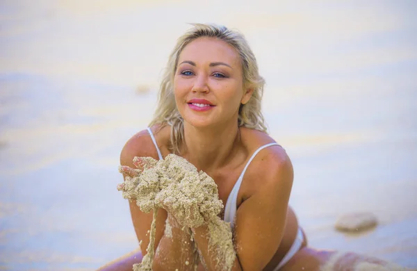 Young beautiful and happy blond woman in white bikini playing with sand cheerful and carefree isolated on tropical paradise desert beach enjoying Summer holidays — Stock Photo, Image