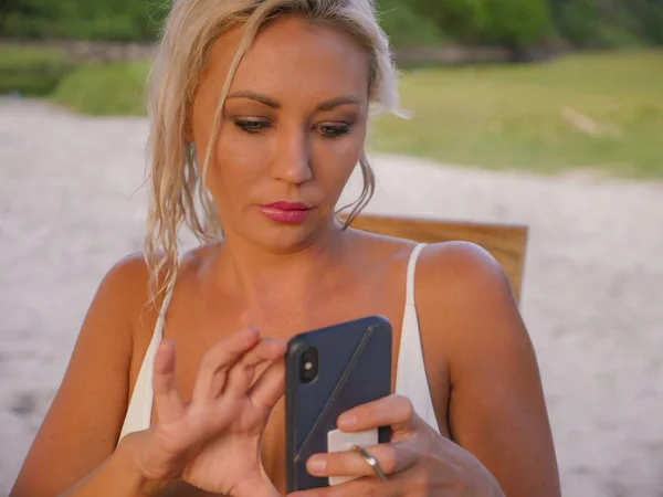 Estilo de vida retrato de la joven atractiva y relajada mujer rubia utilizando el teléfono móvil aplicación de Internet mensajes de texto o el envío de alimentación sentado en el fondo de la playa naturaleza tropical —  Fotos de Stock