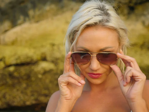 Joven atractiva chica rubia alegre y feliz en bikini sonriente juguetona celebración de gafas de sol posando despreocupado en hermoso fondo de playa disfrutando de vacaciones —  Fotos de Stock