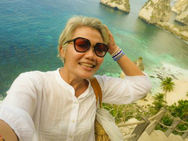 orta yaş 50'lerin mutlu ve neşeli Asyalı kadın gri saçlı güzel tropikal plaj adasında cep telefonu ile selfie alarak yaz zevk uçurumun bakış açısında gülümseyerek