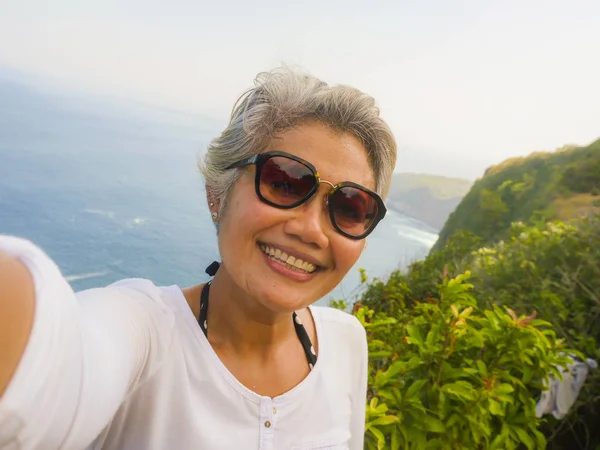 Idade média dos anos 50 feliz e alegre mulher asiática com cabelos grisalhos tirando selfie com telefone celular na bela ilha de praia tropical sorrindo ao ponto de vista do penhasco desfrutando de verão — Fotografia de Stock