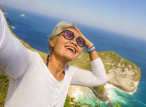 Edad media años 50 feliz y alegre mujer asiática con el pelo gris tomar selfie con el teléfono móvil en la hermosa isla de playa tropical sonriendo en el mirador del acantilado disfrutando de verano — Foto de Stock