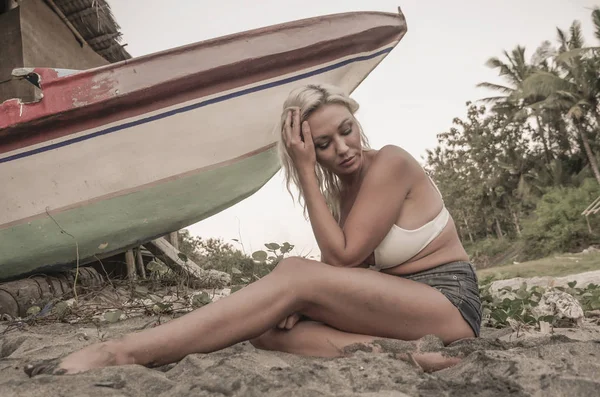 Strand-Lifestyle-Porträt einer jungen attraktiven und entspannten blonden Frau, die am tropischen Strand neben einem alten gestrandeten Boot in einem exotischen Urlaubsreiseziel auf einer Insel sitzt — Stockfoto