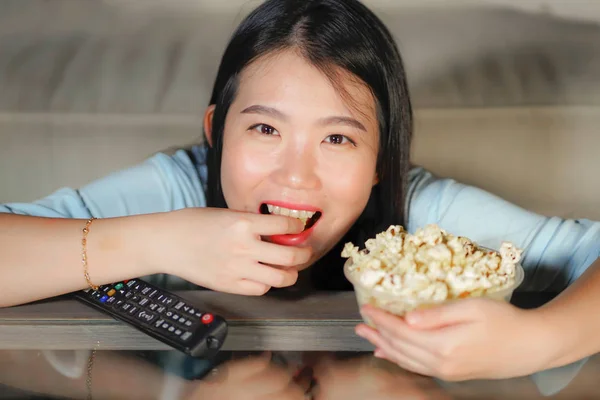 young happy and excited Asian Chinese woman with TV remote controller eating popcorn bowl watching television enjoying Korean drama or comedy movie having fun