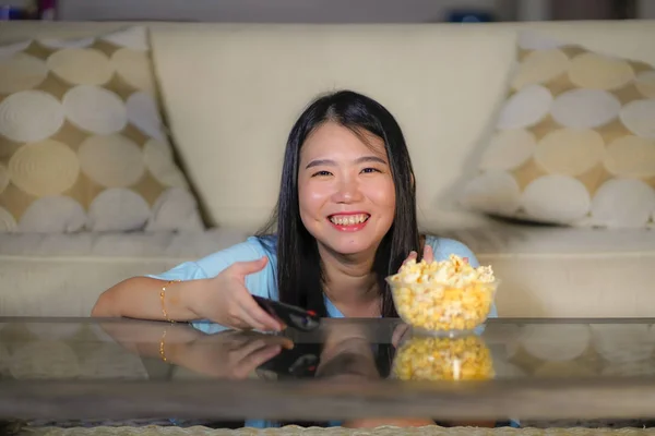 Молодая счастливая и взволнованная азиатская китаянка с пультом дистанционного управления телевизора ест попкорн миску смотреть телевизор наслаждаясь корейской драмы или комедийный фильм весело — стоковое фото