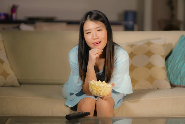 young happy and excited Asian Chinese woman with TV remote controller eating popcorn bowl watching television enjoying Korean drama or comedy movie having fun