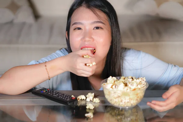 young happy and excited Asian Japanese woman with TV remote controller eating popcorn bowl watching television enjoying Korean drama or comedy movie having fun