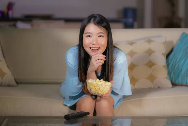 young happy and excited Asian Korean woman with TV remote controller eating popcorn bowl watching television enjoying Korean drama or comedy movie having fun