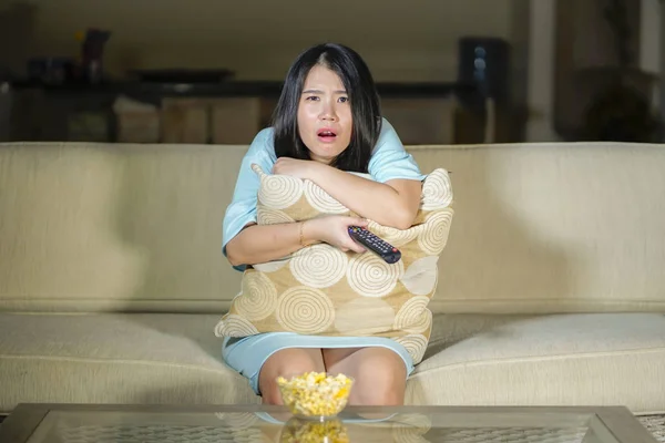 young beautiful and scared Asian Chinese teenager woman in fear watching horror scary movie at home sofa couch eating popcorn bowl holding remote controller