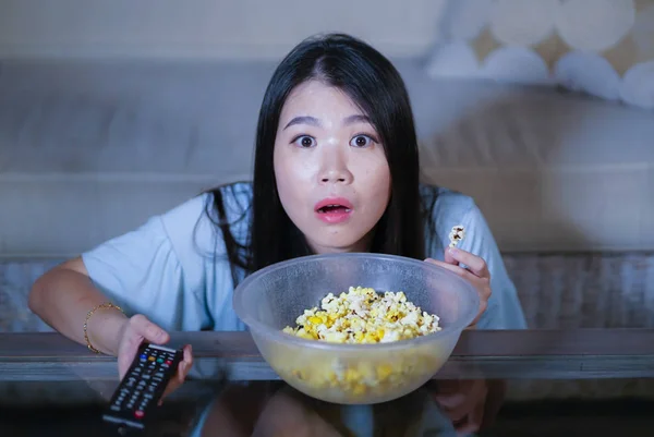 young beautiful and scared Asian Chinese teenager woman in fear watching horror scary movie at home sofa couch eating popcorn bowl holding remote controller