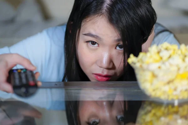 Young beautiful and scared Asian Chinese teenager woman in fear watching horror scary movie at home sofa couch eating popcorn bowl holding remote controller — Stock Photo, Image