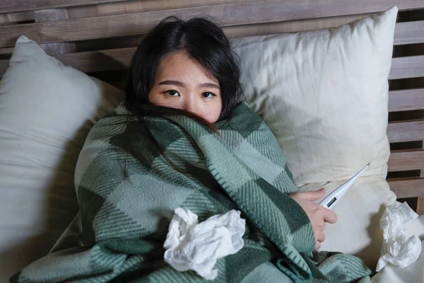 Joven hermosa enferma y agotada mujer china asiática que sufre resfriado y gripe celebración termómetro tener temperatura acostado en la cama cubierta con manta sensación de malestar — Foto de Stock