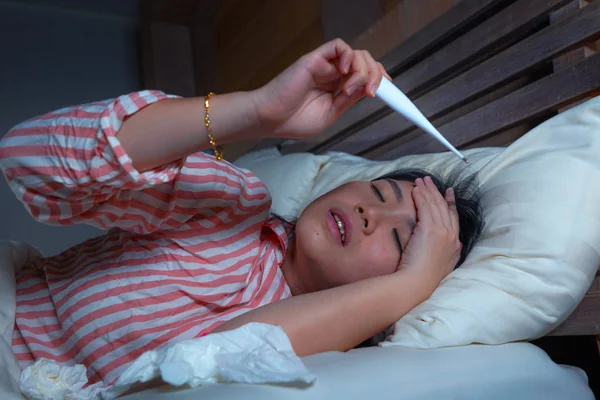 Молодая красивая больная и измученная азиатская китаянка страдает от простуды и гриппа, держа в руках салфетку и термометр, проверяя температуру, лежащую на кровати, чувствуя себя больной и нездоровой — стоковое фото