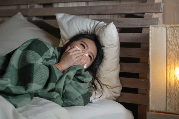 Young beautiful sick and exhausted Asian Japanese woman suffering cold and flu having temperature lying on bed covering with blanket sneezing nose with tissue paper — Stock Photo, Image