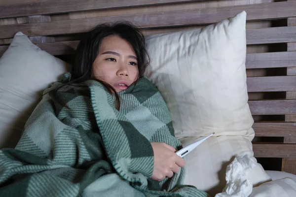 Junge schöne kranke und erschöpfte asiatische Frau, die unter Erkältung und Grippe leidet, hält Thermometer mit Temperatur, die auf der Bettdecke liegt und sich krank fühlt — Stockfoto