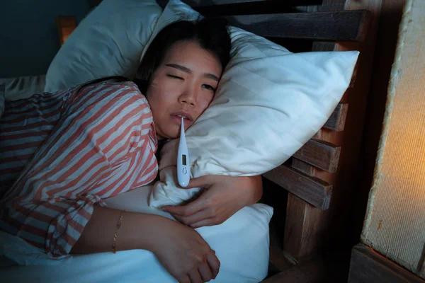 Junge schöne kranke und erschöpfte asiatische Chinesin, die unter Erkältung und Grippe leidet, nimmt Temperatur mit Thermometer, das nachts auf dem Bett liegt und sich krank und unwohl fühlt — Stockfoto