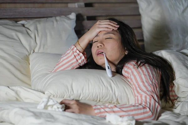 Bela cansado e doente asiático chinês mulher deitado na cama em casa doente sofrendo gripe fria e dor de cabeça sentindo-se mal e febril tomando temperatura com termômetro — Fotografia de Stock