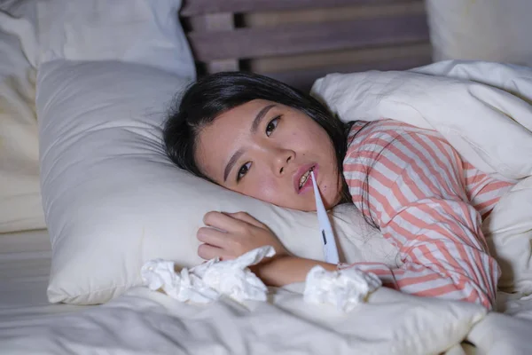 Молодая красивая уставшая и больная азиатская кореянка лежит на кровати дома больная, страдает от простуды и температуры, чувствует себя нездоровой и лихорадочно в медицинской помощи и здоровье — стоковое фото