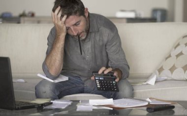 stressed and desperate man at home living room couch doing domestic accounting with paperwork and calculator feeling overwhelmed and worried suffering financial crisis  clipart