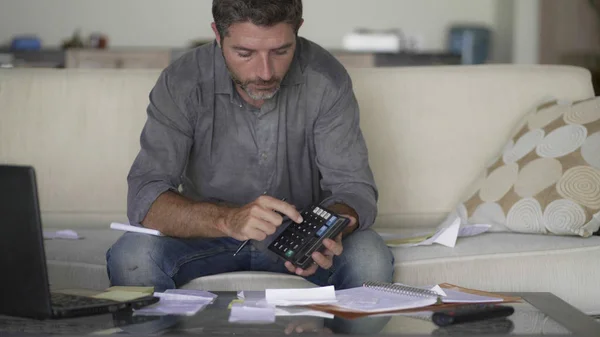 Joven atractivo y ocupado hombre en la sala de estar sofá sofá contabilidad gastos mensuales y pagos de deuda utilizando calculadora contando los ingresos de las finanzas nacionales y los impuestos —  Fotos de Stock