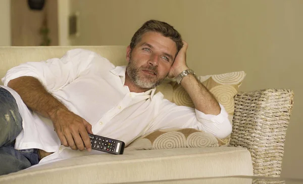 Joven atractivo y feliz hombre viendo programa de televisión o película sosteniendo TV mando a distancia disfrutando relajado tumbado en sofá sofá de la sala de estar en apartamento moderno —  Fotos de Stock