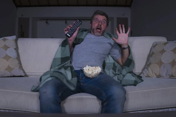 scared and funny man alone at night in living room couch watching horror scary movie in television screaming and eating popcorn covering with blanket